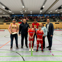 In einer Sporthalle stehen sieben Personen für ein Foto zusammen. Zwei Kinder in roten Trikots und roten Socken befinden sich in der Mitte. Links steht Stadionsprecher Michael Riedl alias Drummy in einem rot-weiß karierten Hemd mit Lederhosenträgern. Zwei